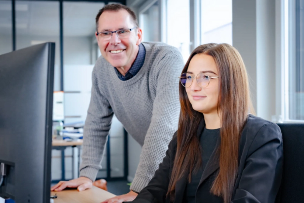 Das Bild zeigt einen älteren Mann und eine jüngere Frau, die vor einem Computer sitzen. Der Mann steht hinter der Frau und legt seine Hände auf den Tisch. Beide lächeln in die Kamera. Es wirkt wie eine Szene aus einem Büro oder einem Arbeitsumfeld, vielleicht ein Mentor und sein Mentee oder Kollegen bei der Arbeit. Die Stimmung ist freundlich und kollegial.