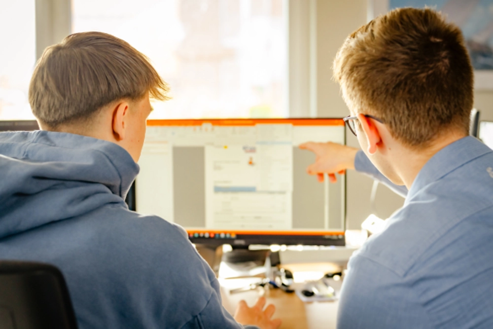 Nahaufnahme von zwei jungen Männern, die an einem Schreibtisch sitzen und gemeinsam an einem Computer arbeiten. Der Mann auf der linken Seite trägt einen blauen Kapuzenpulli, sein Rücken ist der Kamera zugewandt, und nur sein Hinterkopf und seine Schultern sind zu sehen. Er scheint aufmerksam zuzuhören. Der Mann auf der rechten Seite, dessen Gesicht teilweise sichtbar ist, trägt eine Brille und ein hellblaues Hemd mit Kragen. Er gestikuliert in Richtung des Computerbildschirms und scheint der anderen Person etwas zu erklären.