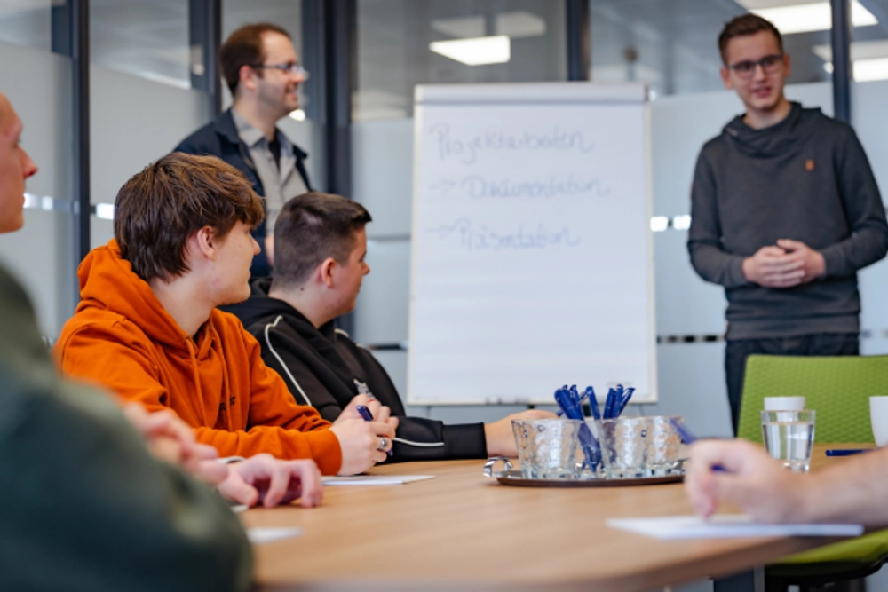 Das Foto zeigt eine Gruppe junger Erwachsener, die um einen hellbraunen Holztisch in einem modernen Büro sitzen.