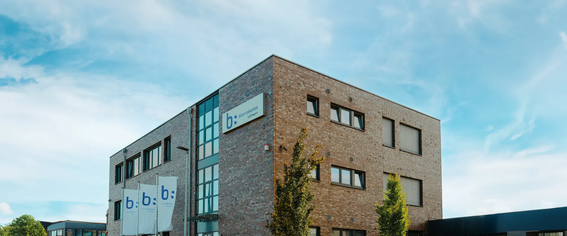 Das Bild zeigt ein modernes, dreistöckiges Bürogebäude aus Backstein unter einem leicht bewölkten Himmel. Das Gebäude trägt das Logo „b: blue-solution software“. Vor dem Gebäude stehen einige Fahnen mit demselben Logo. Die Architektur ist schlicht und funktional.